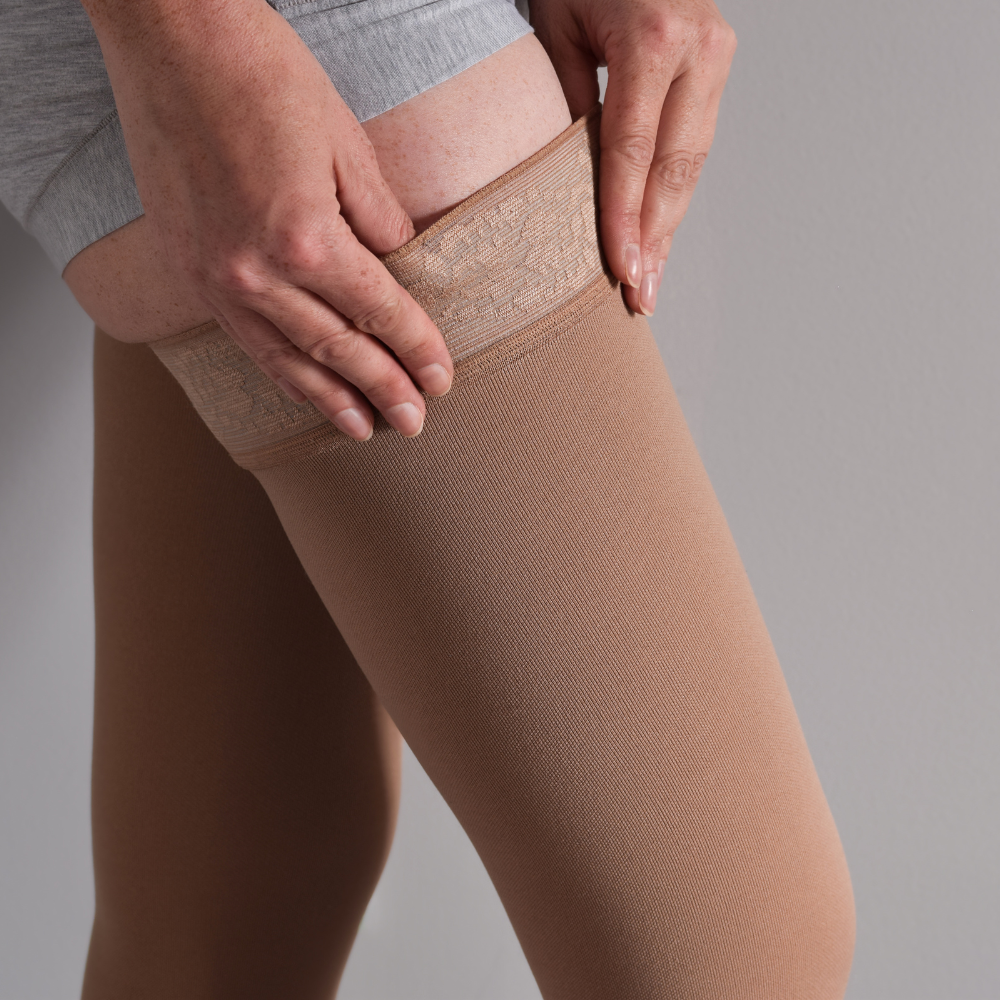 Nahaufnahme von Händen, die einen hautfarbenen Kompressionsstrumpf am Oberschenkel nach oben ziehen, um die Blutzirkulation zu unterstützen oder Schwellungen vorzubeugen.