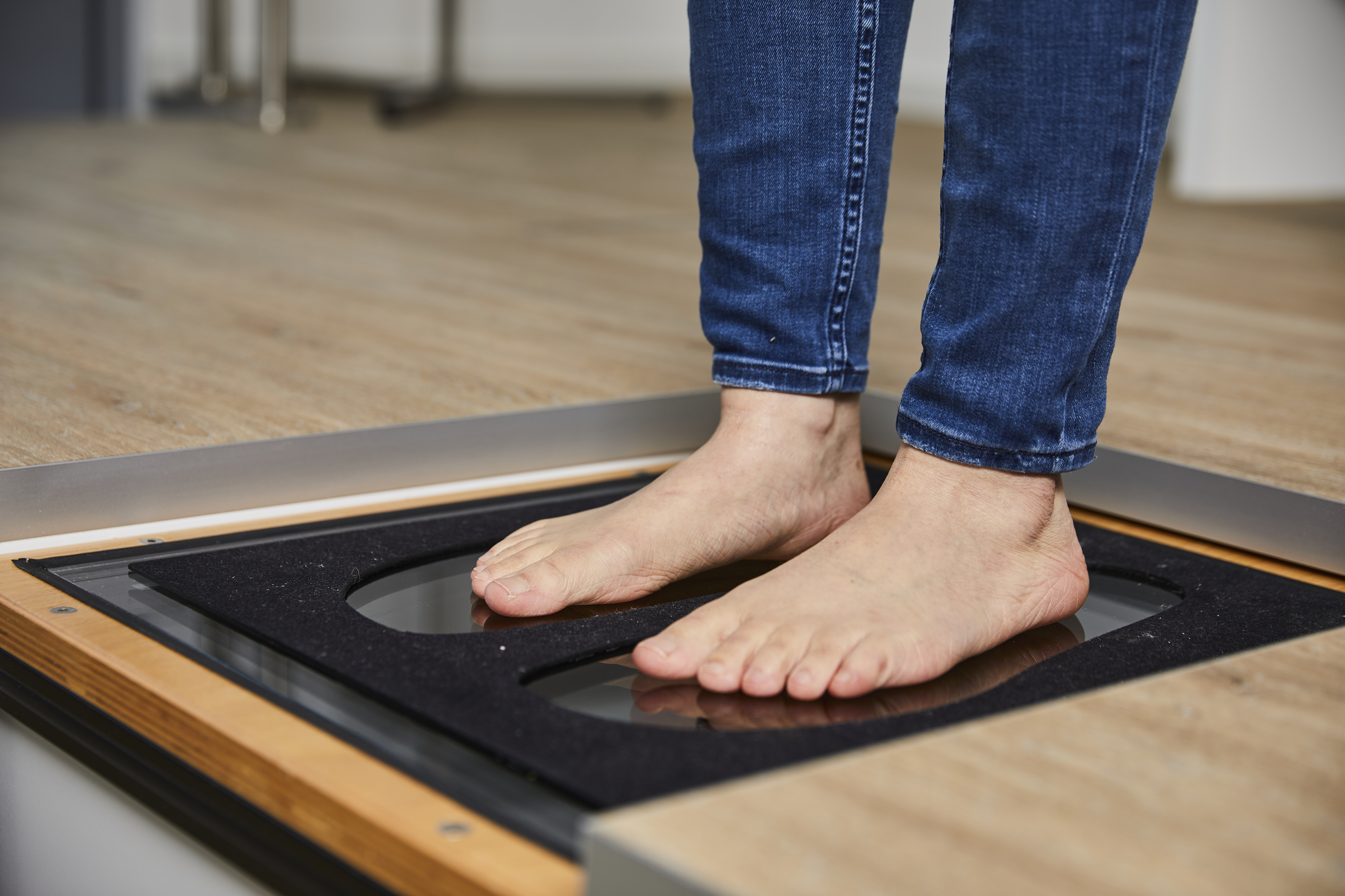 Eine Person steht barfuß auf einem Gerät zur 3D-Fußanalyse. Das Gerät misst die Fußform und den Druckverlauf, während die Person auf der Glasplatte steht.