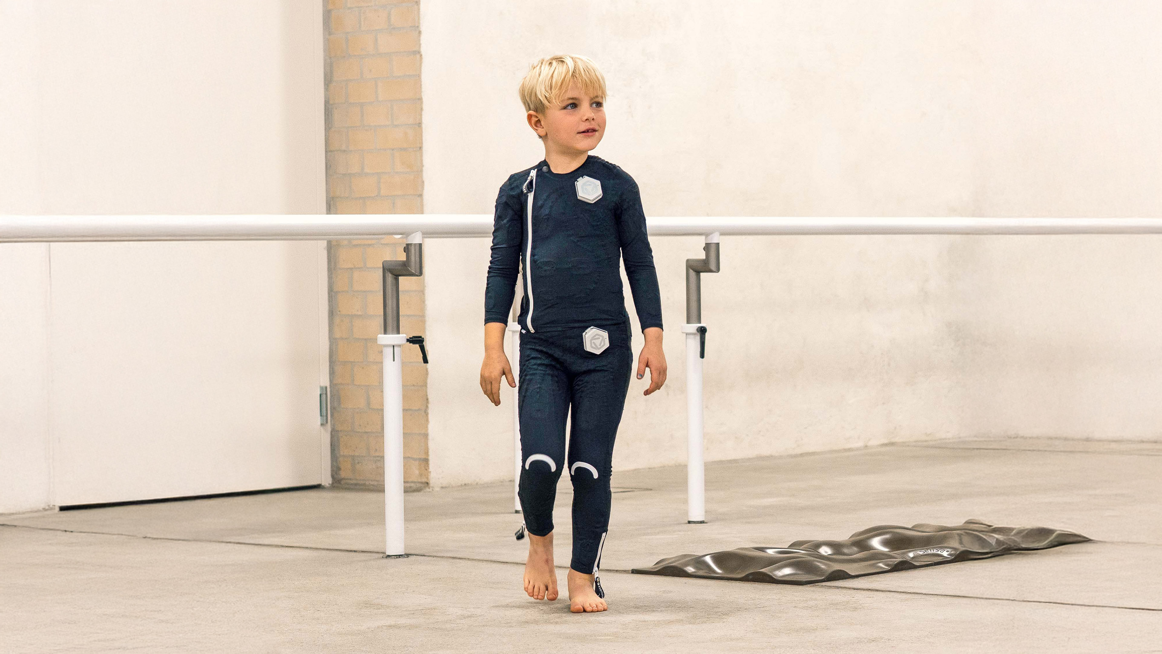 kleiner Junge mit blonden Haaren im exopulse suit für Kinder vor einem Barren